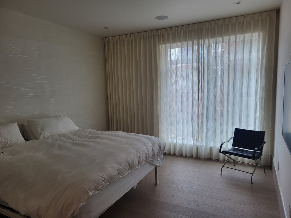 A serene master bedroom featuring cream and white bedding, motorized drapery shades in neutral tones, and