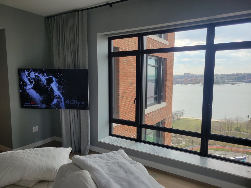 Contemporary bedroom with TV mounted on deep blue accent wall. Gray drapery frames the windows. Bed
