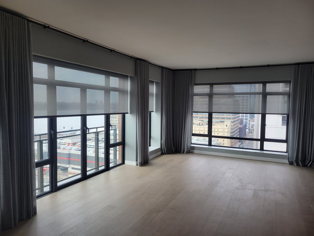 Modern living room with extensive window walls featuring roller shades and full-length drapery in charcoal